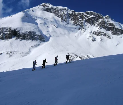 Vier Wanderer steigen im Schnee vor schneebedecktem Berg unter blauem Himmel auf
