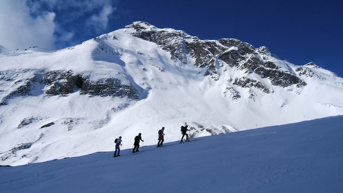 Vier Wanderer steigen im Schnee vor schneebedecktem Berg unter blauem Himmel auf