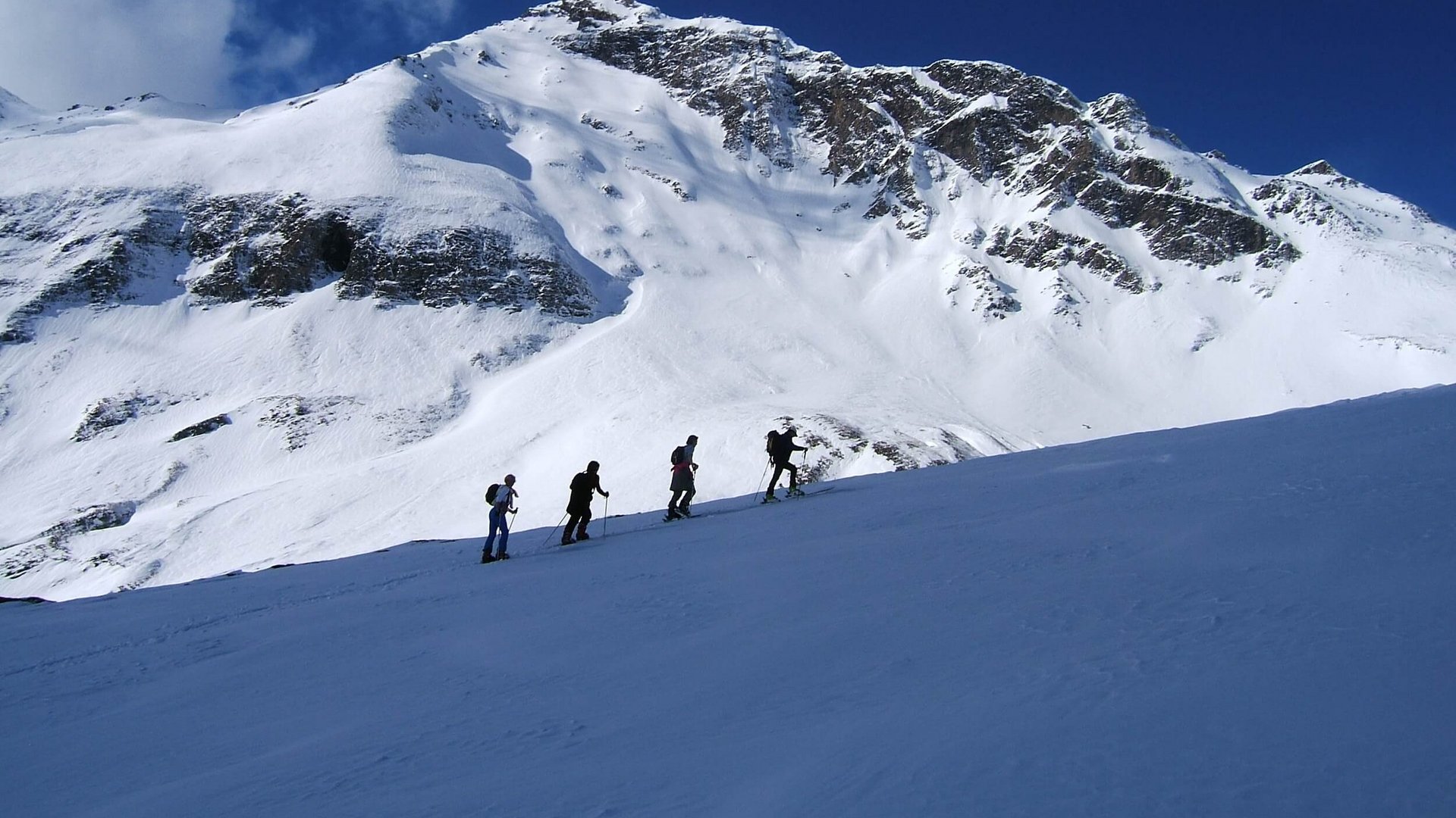 Four hikers ascending snowy slope below snow-covered mountain and clear blue sky
