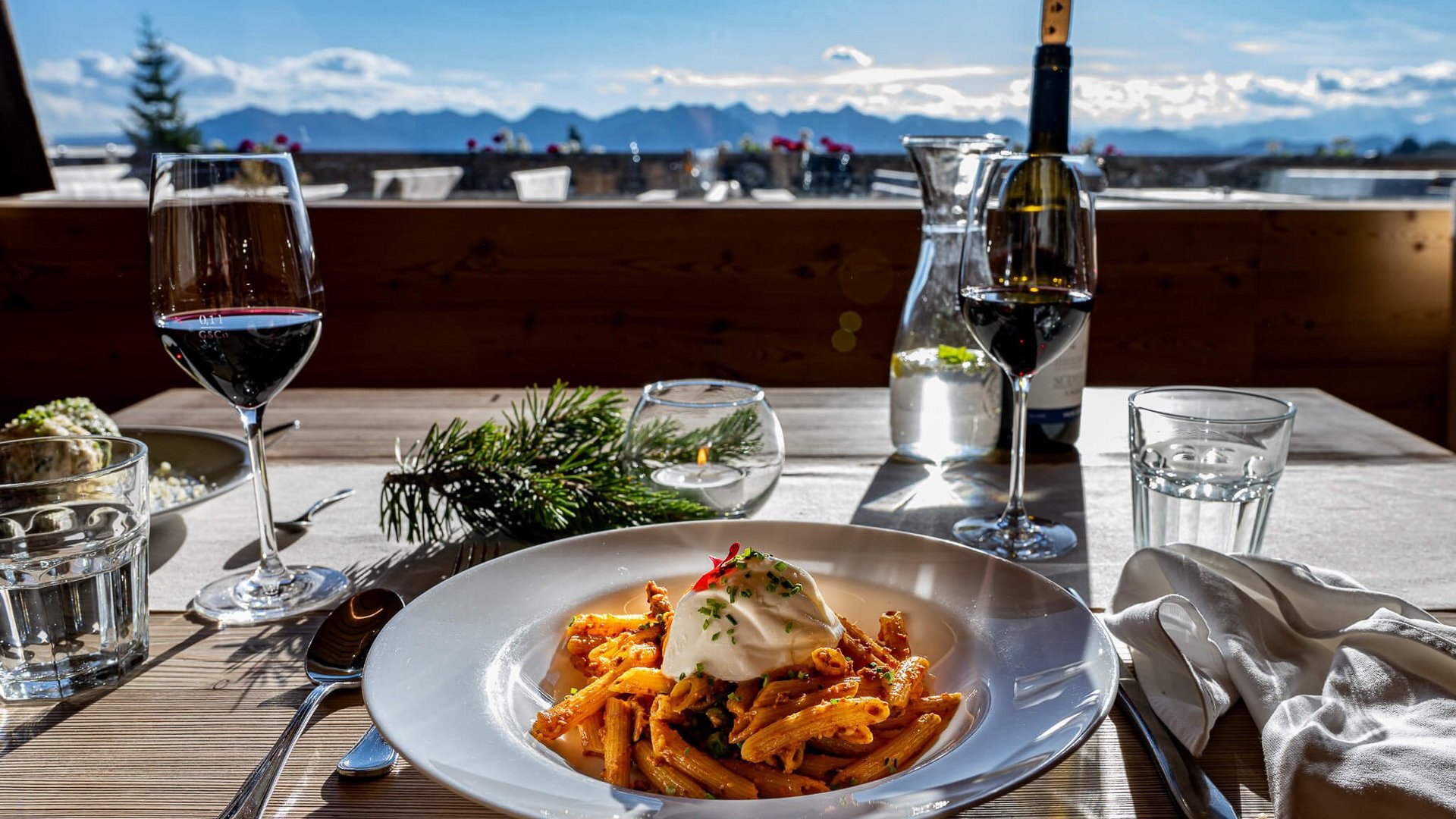 Pasta mit Sahne auf einem Teller, Rotwein und Wasserglas auf Holztisch mit Bergblick