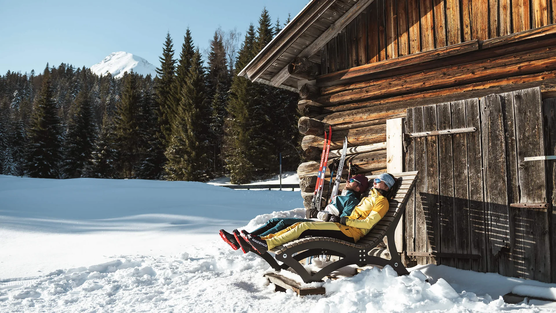 Zwei Personen entspannen auf Liegestühlen neben einer Holzhütte im Schnee
