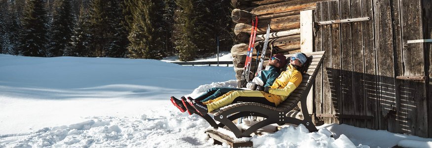 Zwei Personen entspannen auf Liegestühlen neben einer Holzhütte im Schnee