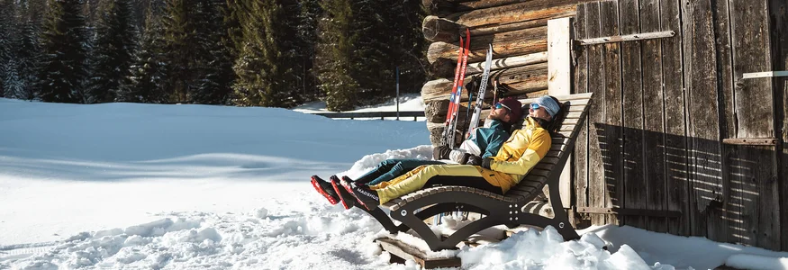 Zwei Personen entspannen auf Liegestühlen neben einer Holzhütte im Schnee