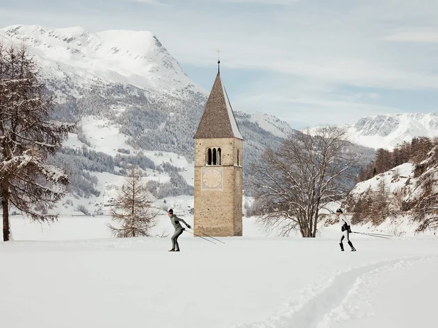 Two cross-country skiers near a church tower on snowy plain with mountains
