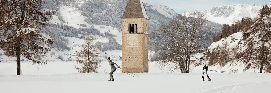 Zwei Skilangläufer vor einem Kirchturm auf einer schneebedeckten Ebene mit Bergen