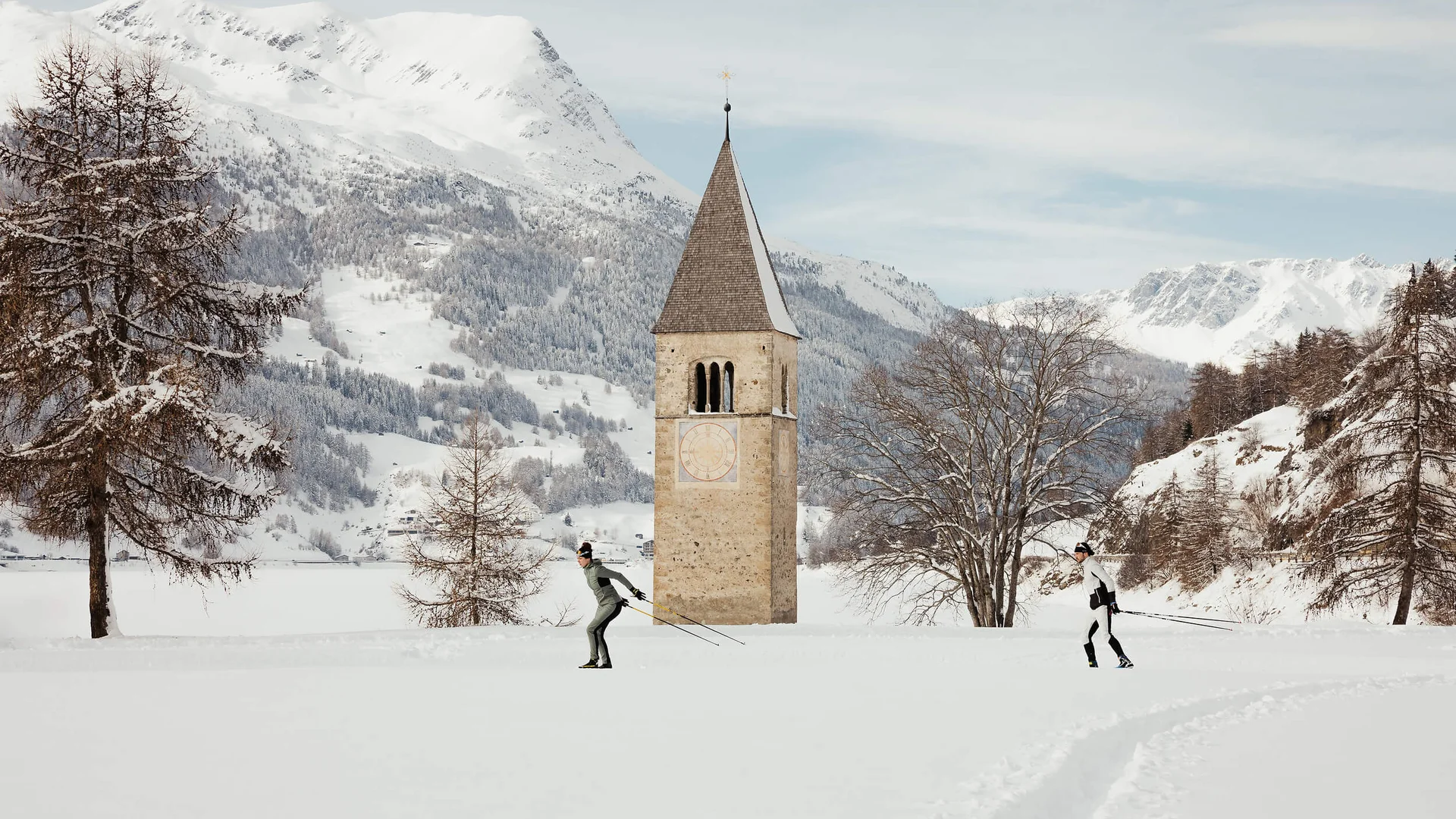 Zwei Skilangläufer vor einem Kirchturm auf einer schneebedeckten Ebene mit Bergen