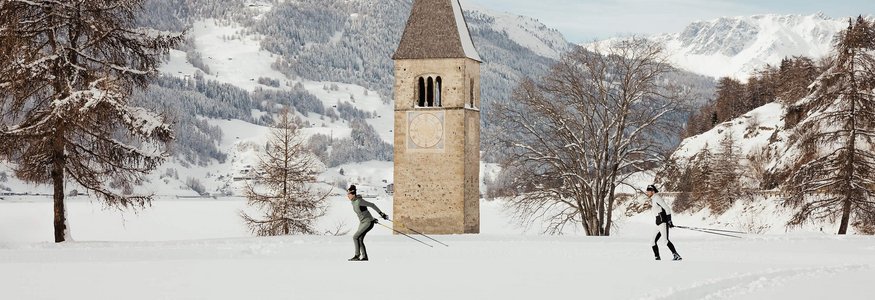 Zwei Skilangläufer vor einem Kirchturm auf einer schneebedeckten Ebene mit Bergen