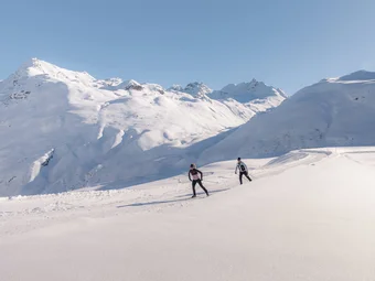 Two cross-country skiers in snowy mountain landscape under clear sky