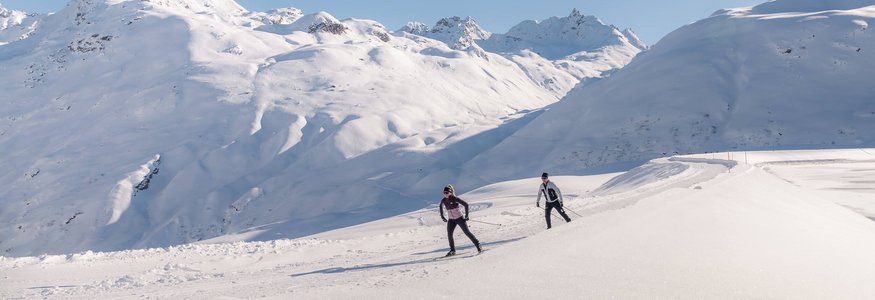 Two cross-country skiers in snowy mountain landscape under clear sky