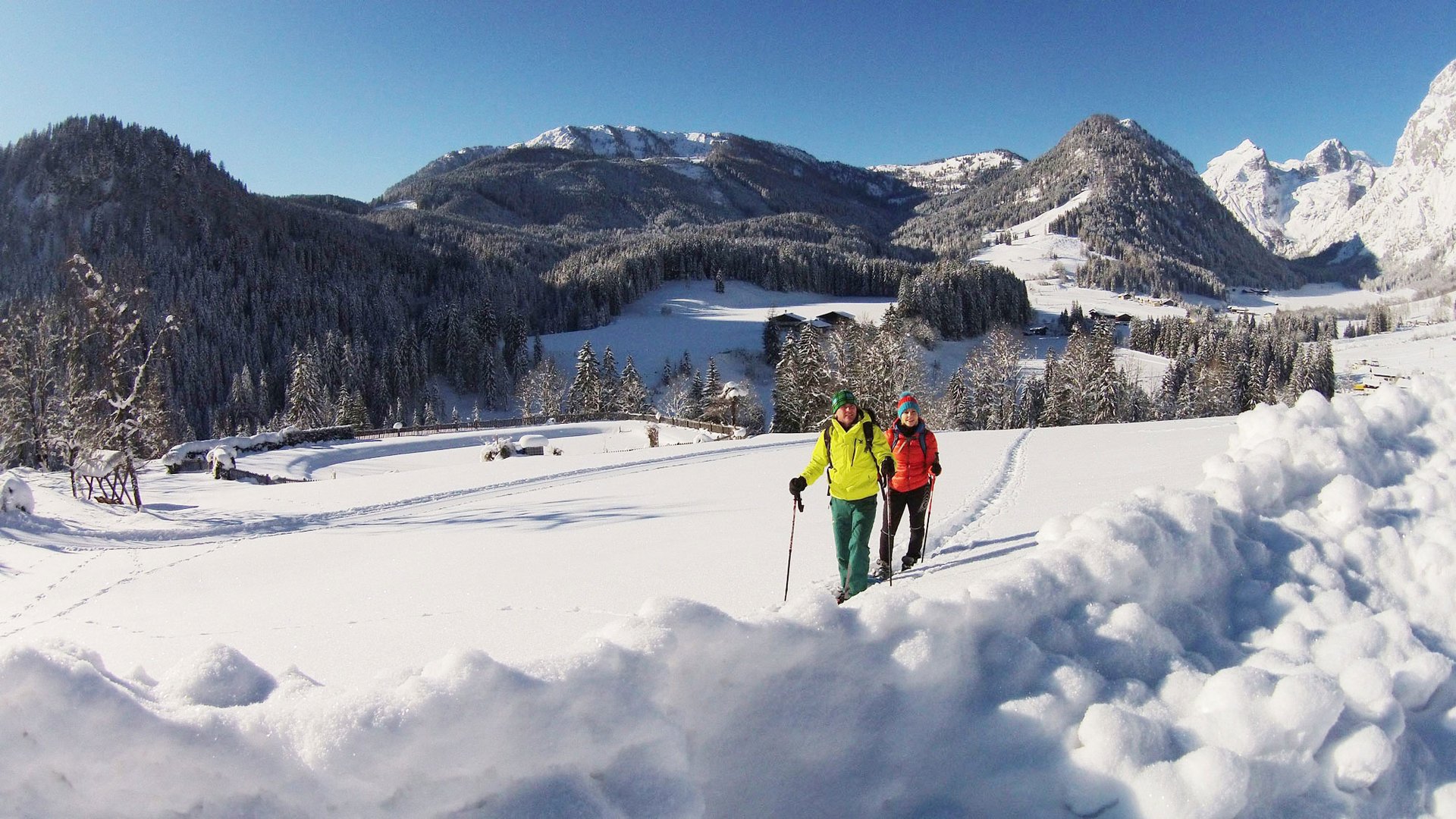 Zwei Personen wandern im Schnee mit Bergen und Wald im Hintergrund