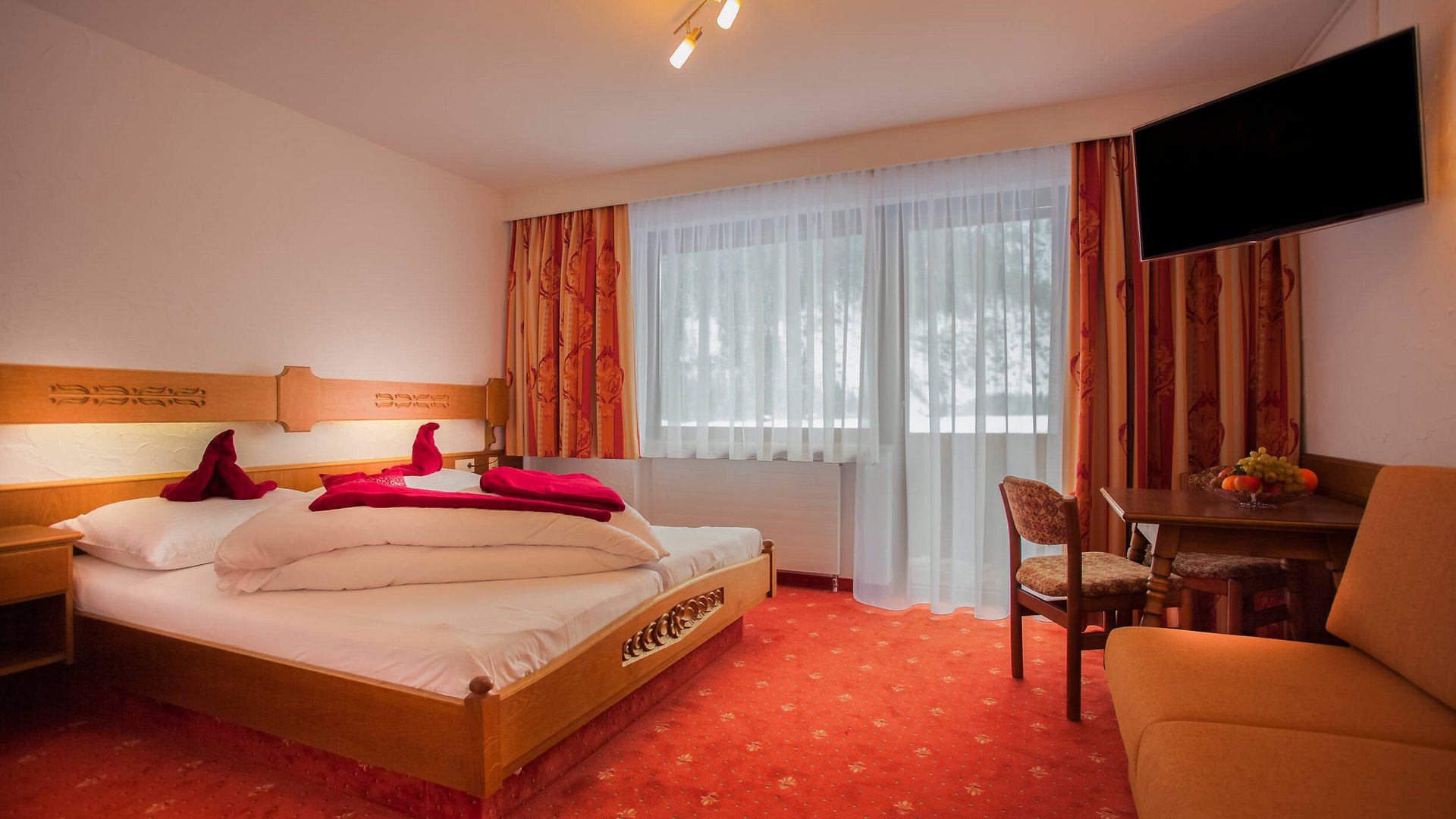 Bright hotel room with wooden bed, red carpet, and wall-mounted TV