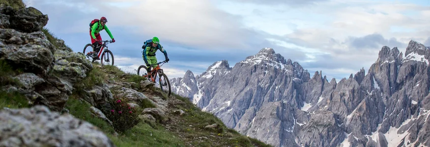 Fit durch den Sommer © Markus Greber Zwei Mountainbiker fahren an einem steilen Berg mit schneebedeckten Gipfeln im Hintergrund