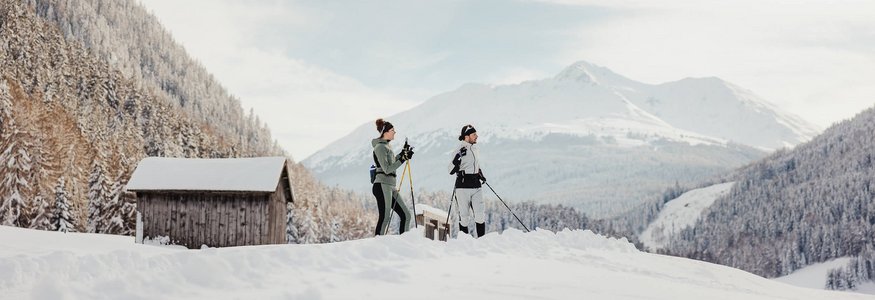 Zwei Personen beim Langlaufen im verschneiten Bergtal mit Hütten im Hintergrund