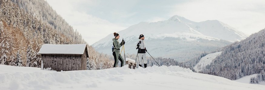Zwei Personen beim Langlaufen im verschneiten Bergtal mit Hütten im Hintergrund