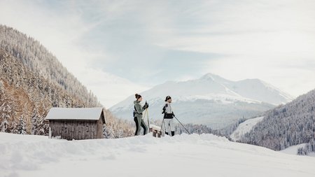 Zwei Personen beim Langlaufen im verschneiten Bergtal mit Hütten im Hintergrund