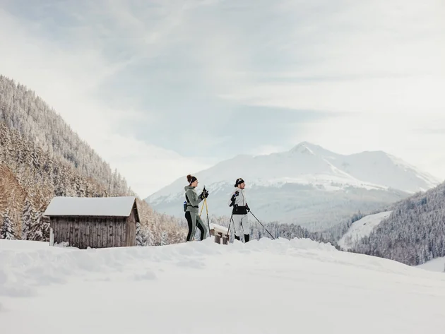 Two people cross-country skiing in snowy mountain valley with huts in background