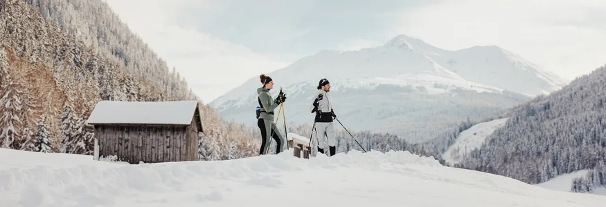 Zwei Personen beim Langlaufen im verschneiten Bergtal mit Hütten im Hintergrund
