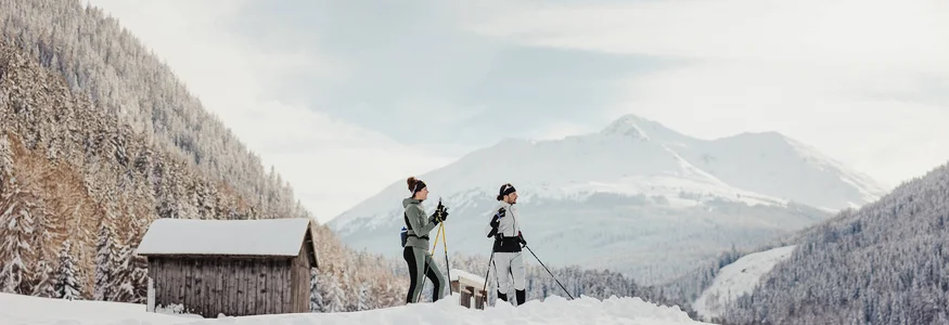 Zwei Personen beim Langlaufen im verschneiten Bergtal mit Hütten im Hintergrund