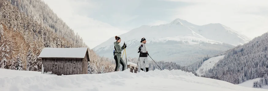 Zwei Personen beim Langlaufen im verschneiten Bergtal mit Hütten im Hintergrund