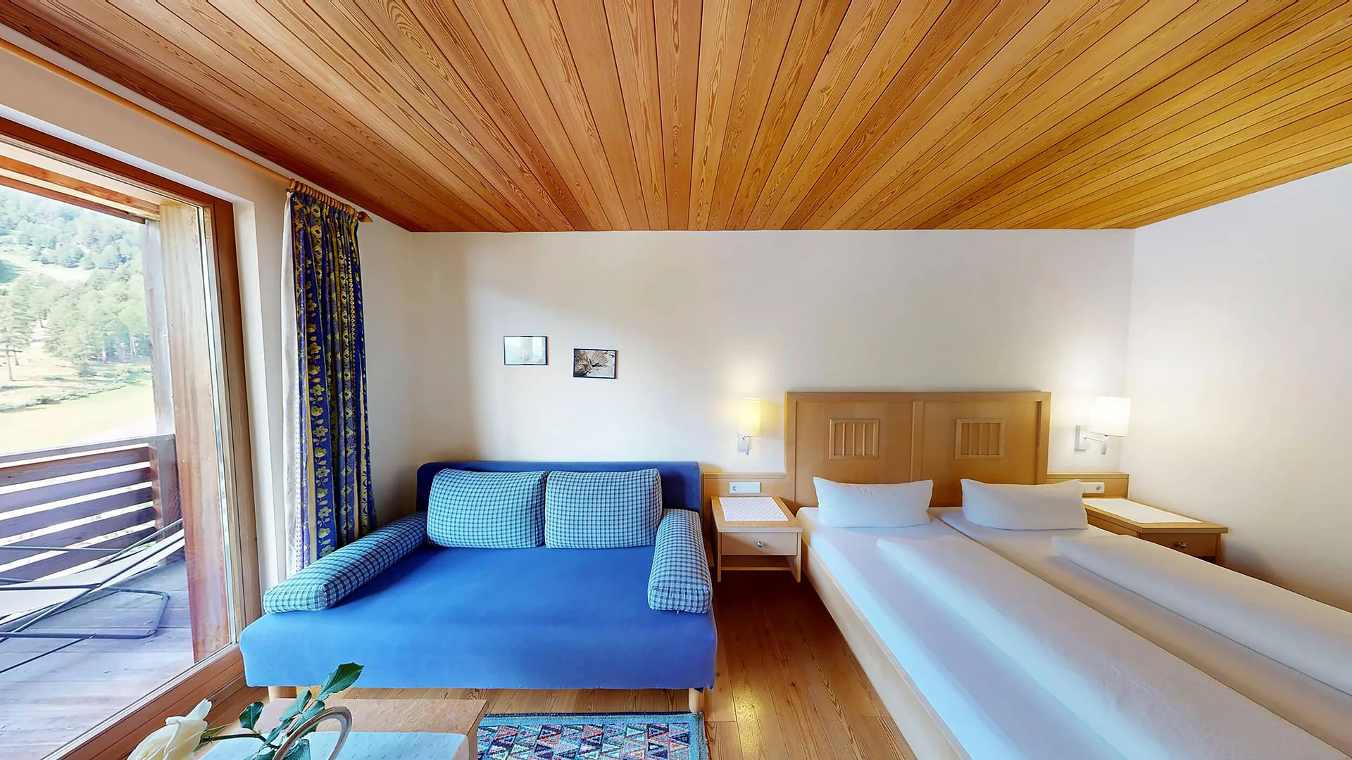 Helles Hotelzimmer mit zwei Betten, blauem Sofa und Balkon mit Bergblick