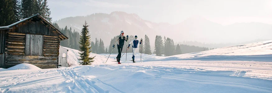 Kleinwalsertal © Kleinwalsertal Tourismus - Oliver Farys Zwei Langläufer neben einer Holzhütte in verschneiter Berglandschaft