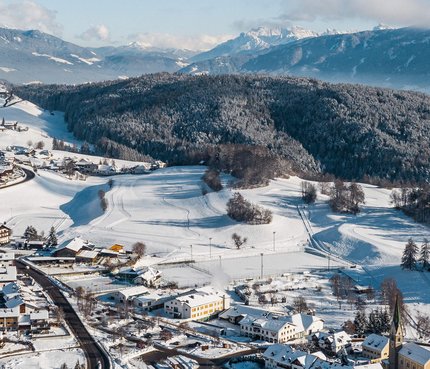 Verschneites Dorf zwischen bewaldeten Hügeln und Bergen im Hintergrund