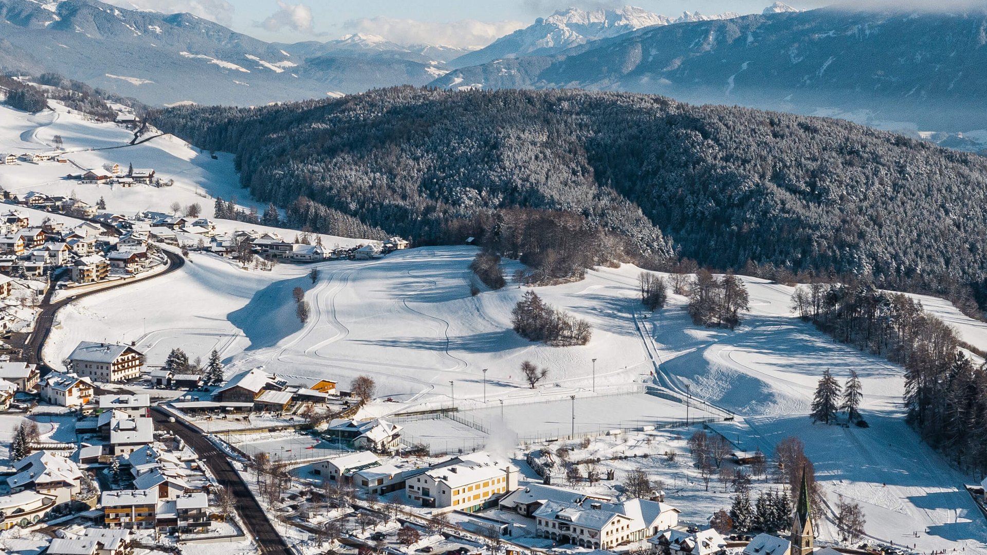 Verschneites Dorf zwischen bewaldeten Hügeln und Bergen im Hintergrund
