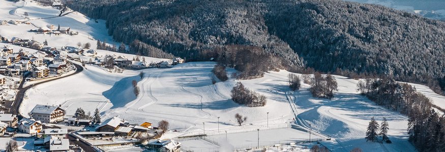 Verschneites Dorf zwischen bewaldeten Hügeln und Bergen im Hintergrund