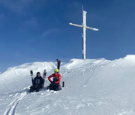 Zwei Skifahrer sitzen im Schnee unter einem großen vereisten Gipfelkreuz