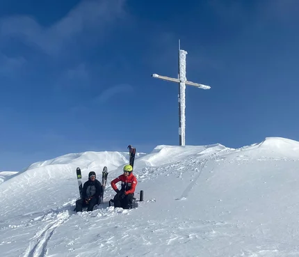 Two skiers sit in snow below a large icy summit cross