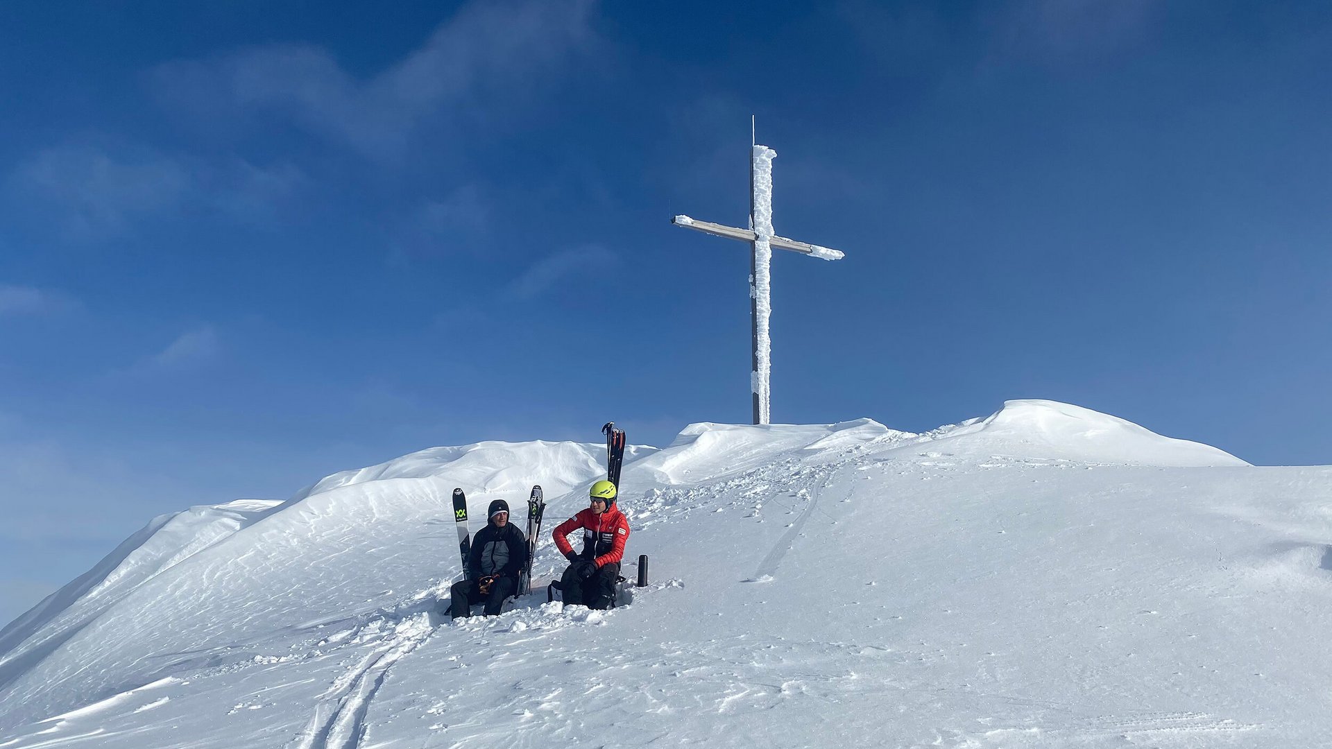 Two skiers sit in snow below a large icy summit cross