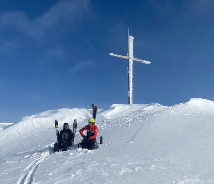Two skiers sit in snow below a large icy summit cross