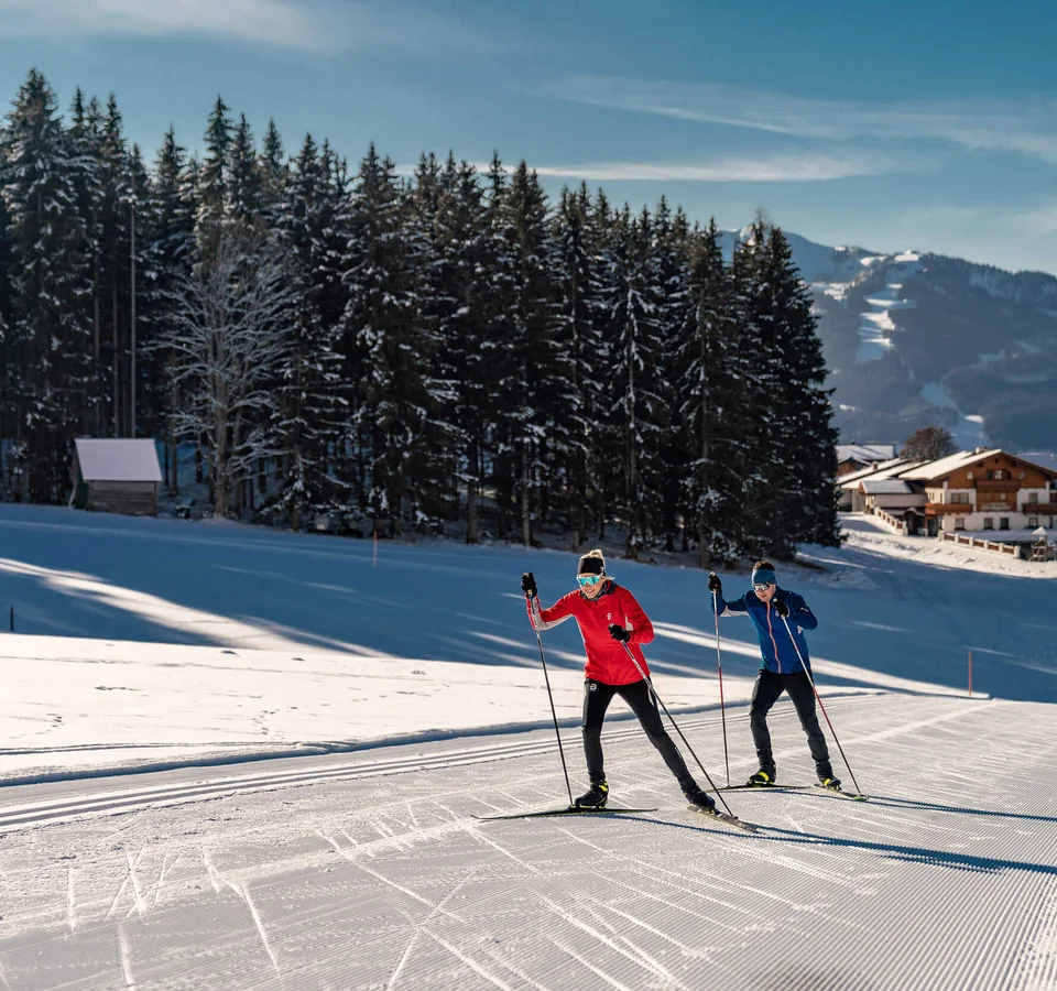 Two cross-country skiers on snow trail with forest and mountains