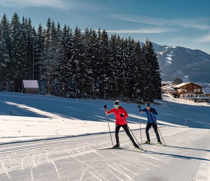Zwei Langläufer auf verschneiter Loipe vor Wald und Bergen
