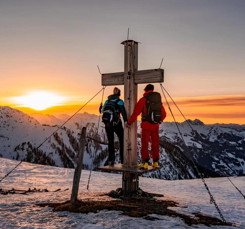 Zwei Wanderer stehen an einem Gipfelkreuz bei Sonnenuntergang in verschneiten Bergen