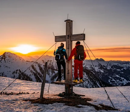 Zwei Wanderer stehen an einem Gipfelkreuz bei Sonnenuntergang in verschneiten Bergen