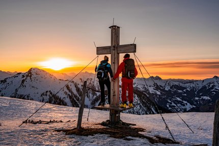 Zwei Wanderer stehen an einem Gipfelkreuz bei Sonnenuntergang in verschneiten Bergen