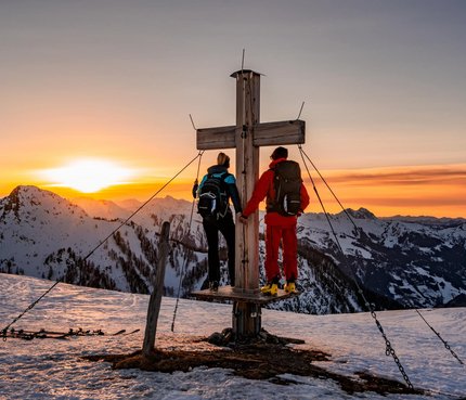 Two hikers standing at a summit cross during sunset in snowy mountains