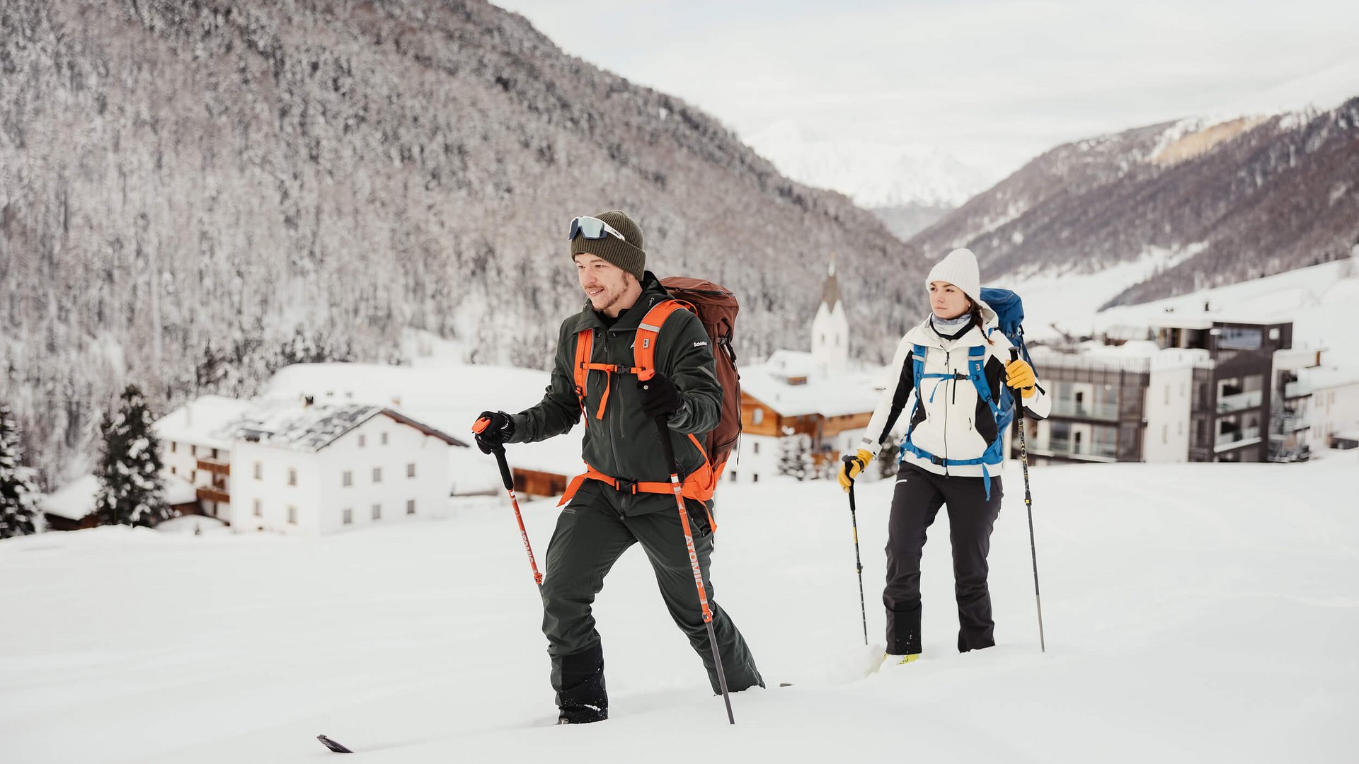Zwei Skitourengeher mit Rucksäcken und Stöcken im verschneiten Bergdorf