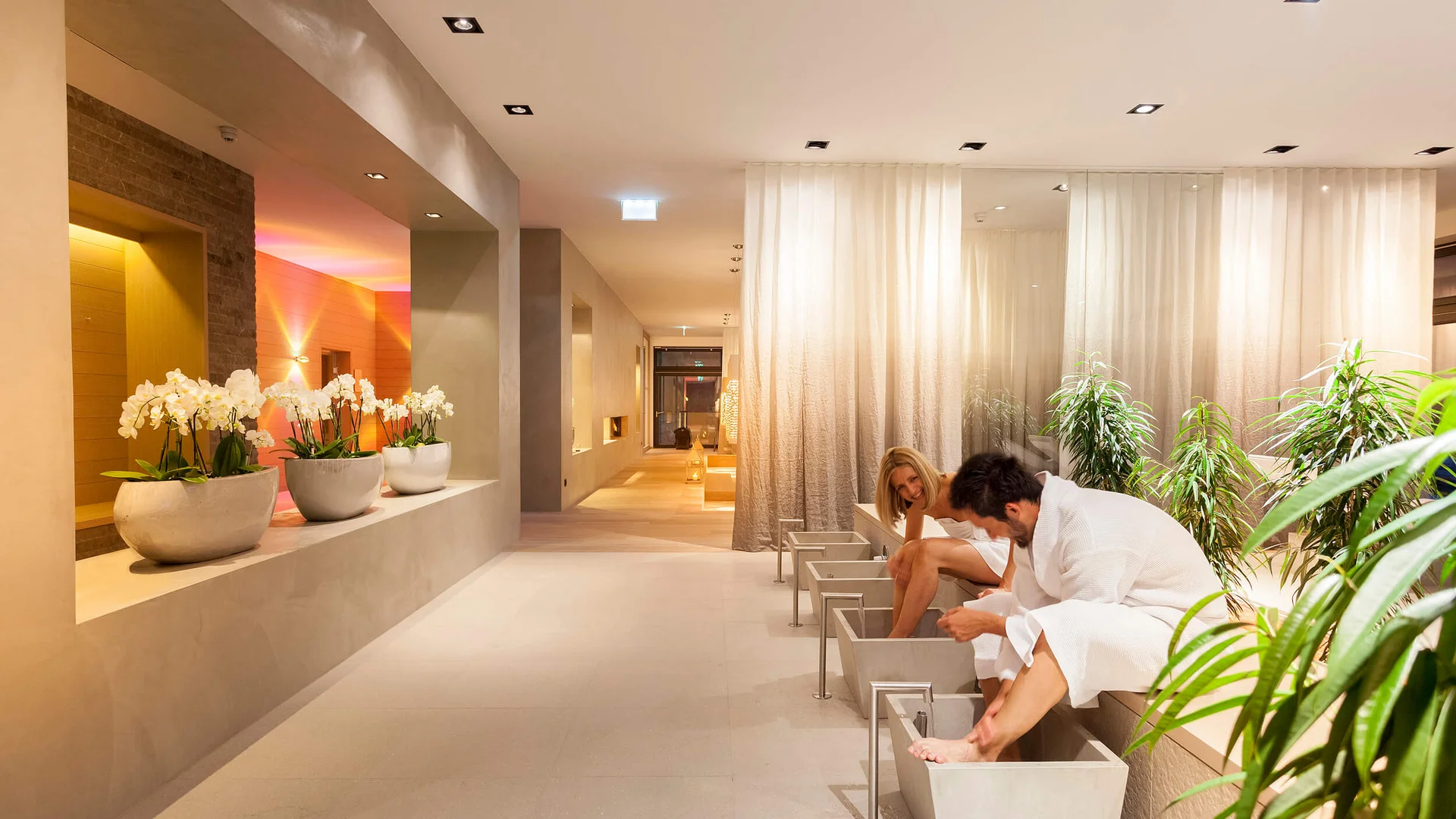Spa-Bereich mit zwei Personen in weißen Bademänteln bei einem Fußbad, Orchideen und Pflanzen
