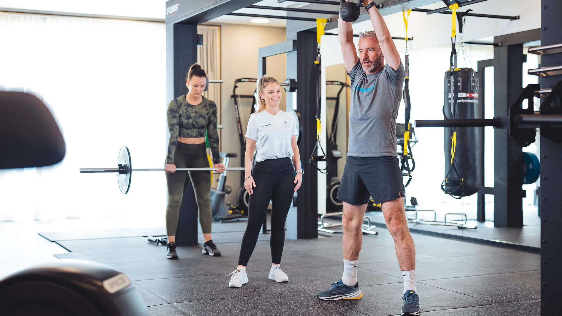 Mann hebt Kettlebell im Fitnessstudio, Frau mit Langhantel, Trainerin beobachtet