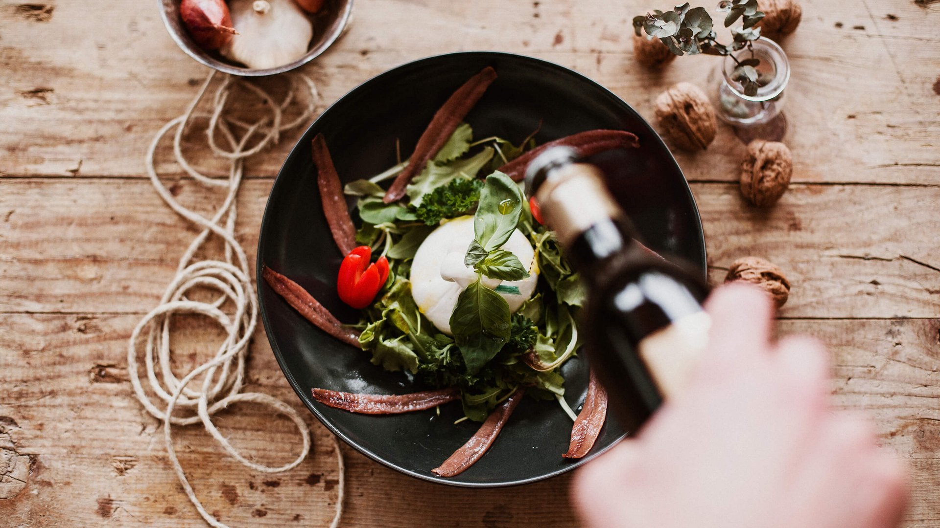 Hand gießt Öl über Burrata mit Tomate und Sardellen auf einem Holzbrett