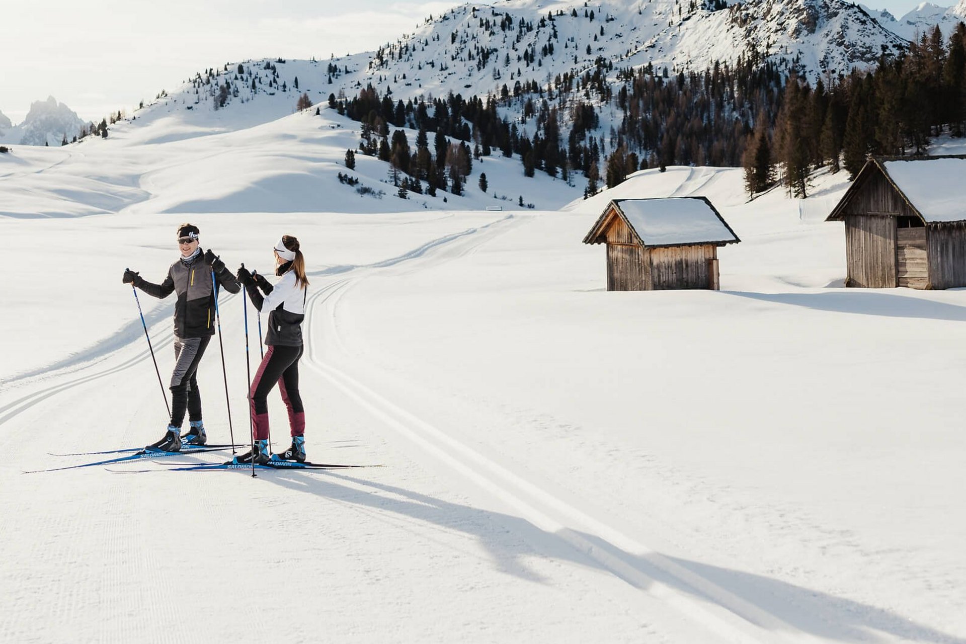 Two cross-country skiers on snowy trail near cabins and mountains