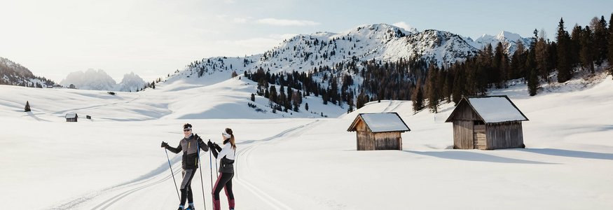Zwei Langläufer auf verschneiter Loipe mit Hütten und Bergen im Hintergrund