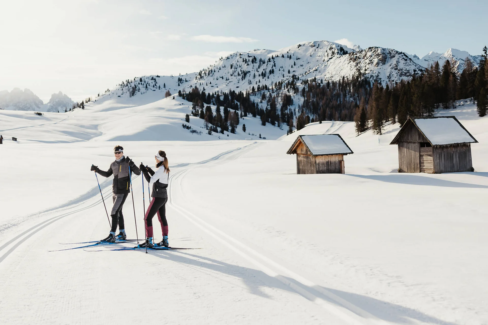 Zwei Langläufer auf verschneiter Loipe mit Hütten und Bergen im Hintergrund