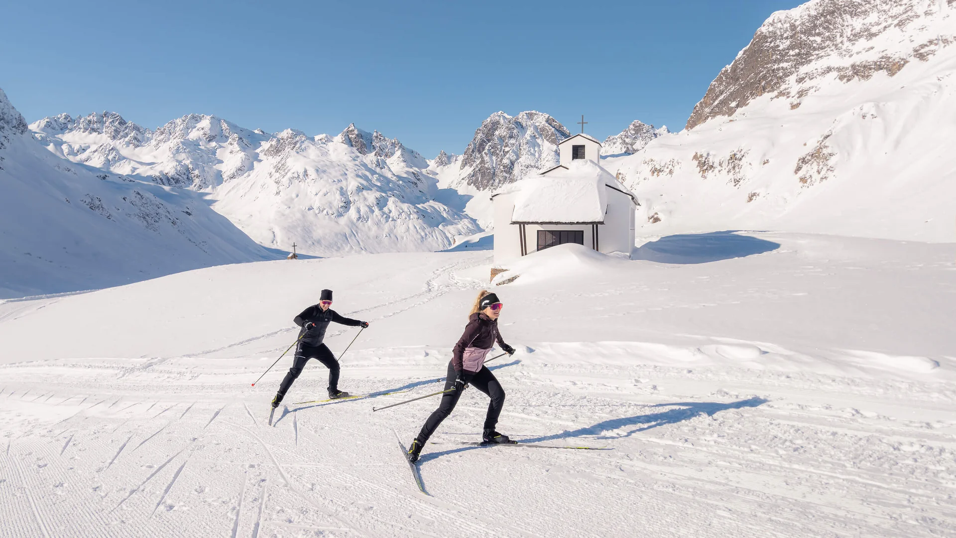 Loipen & Loipenbericht © TVB Paznaun-Ischgl Zwei Skilangläufer vor verschneiter Kapelle in den Bergen unter klarem Himmel