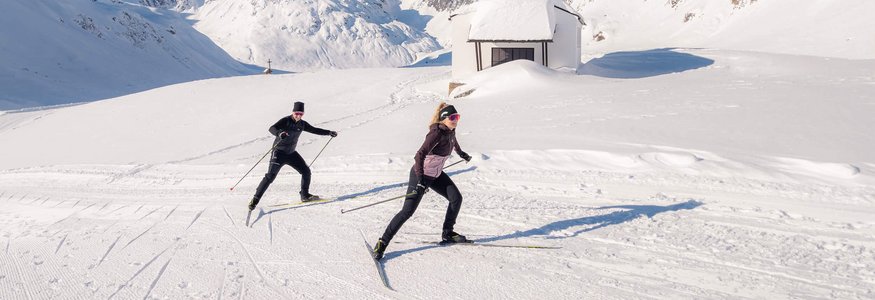 Zwei Skilangläufer vor verschneiter Kapelle in den Bergen unter klarem Himmel