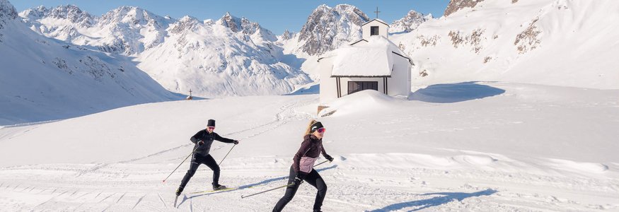 Zwei Skilangläufer vor verschneiter Kapelle in den Bergen unter klarem Himmel
