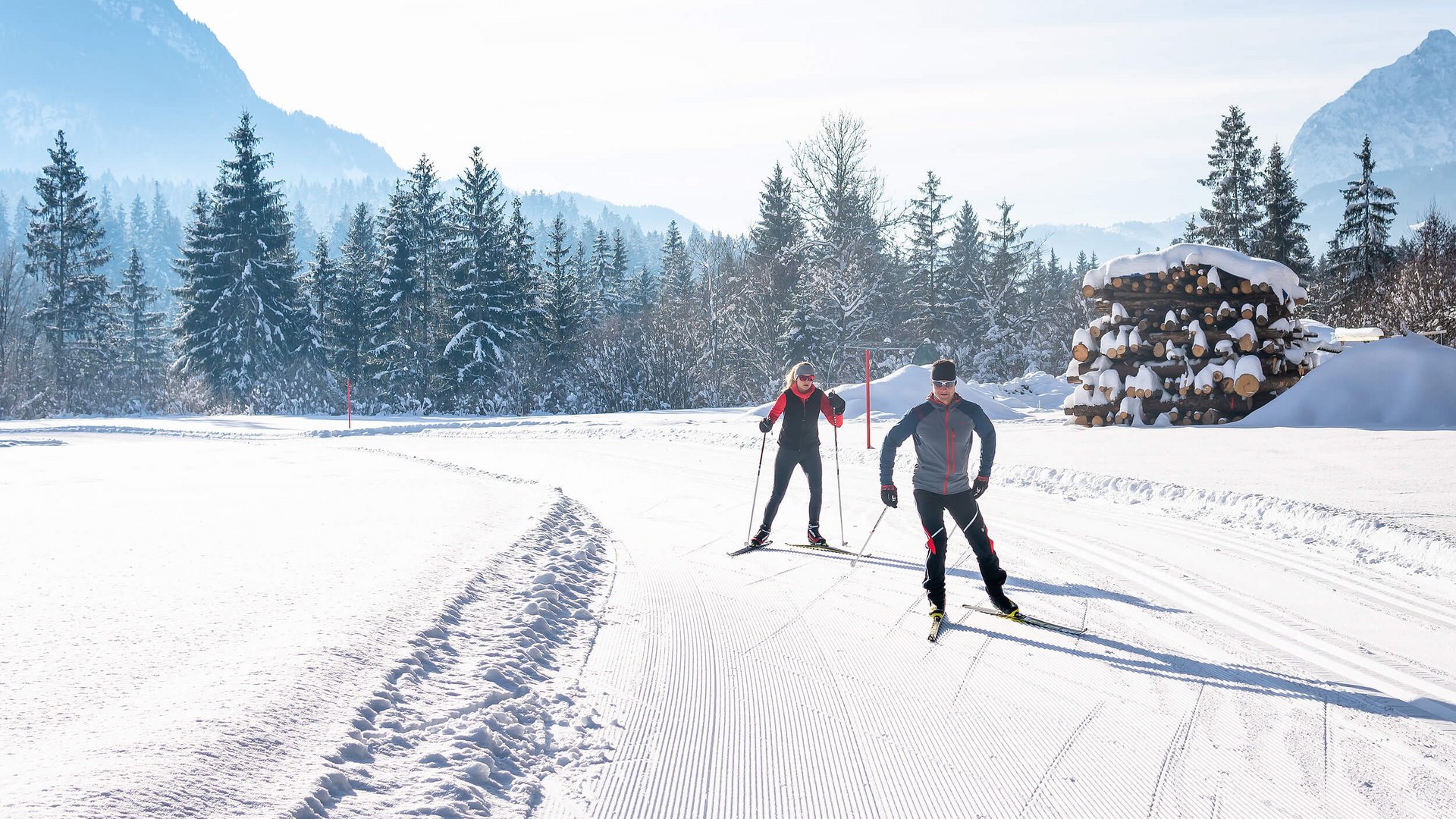Zwei Langläufer auf schneebedeckter Loipe vor verschneiten Bergen