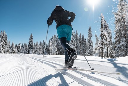 Person beim Langlaufen auf sonniger, verschneiter Strecke mit blauen Himmel